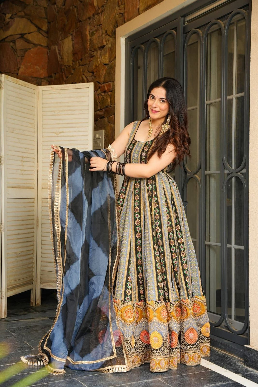 Woman in traditional Anarkali Gown holding a blue and black patterned scarf, standing in front of a stone wall and metal gate.