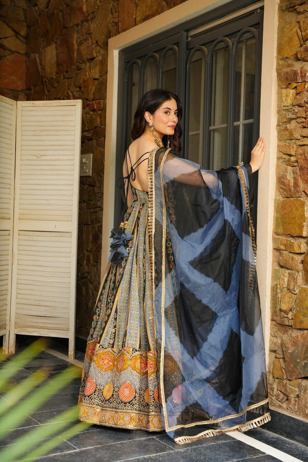 Woman in a traditional Anarkali Gown with a blue and black patterned shawl, standing in a doorway.