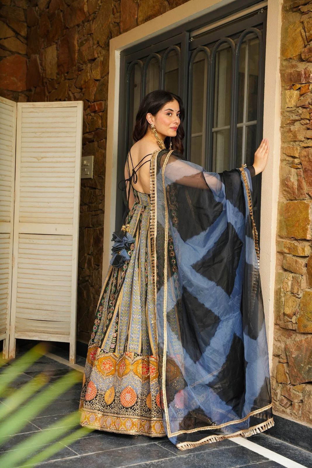 Woman in a traditional Anarkali Gown with a blue and black patterned shawl, standing in a doorway.