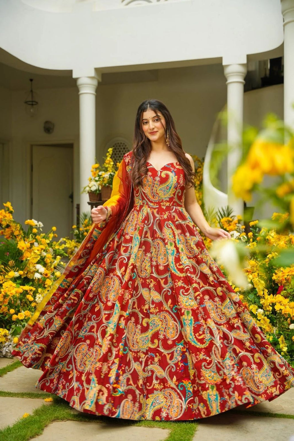 Woman in a red and gold traditional Anarkali Gown standing in a garden with flowers.