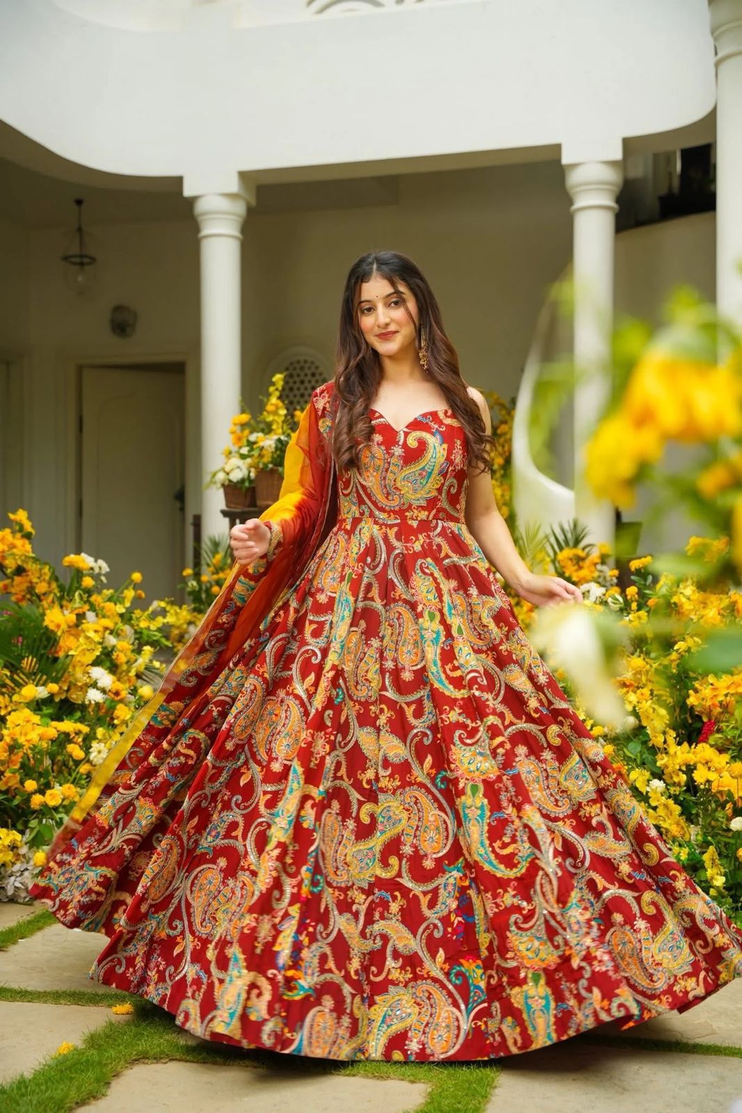 Woman in a red and gold traditional Anarkali Gown standing in a garden with flowers.