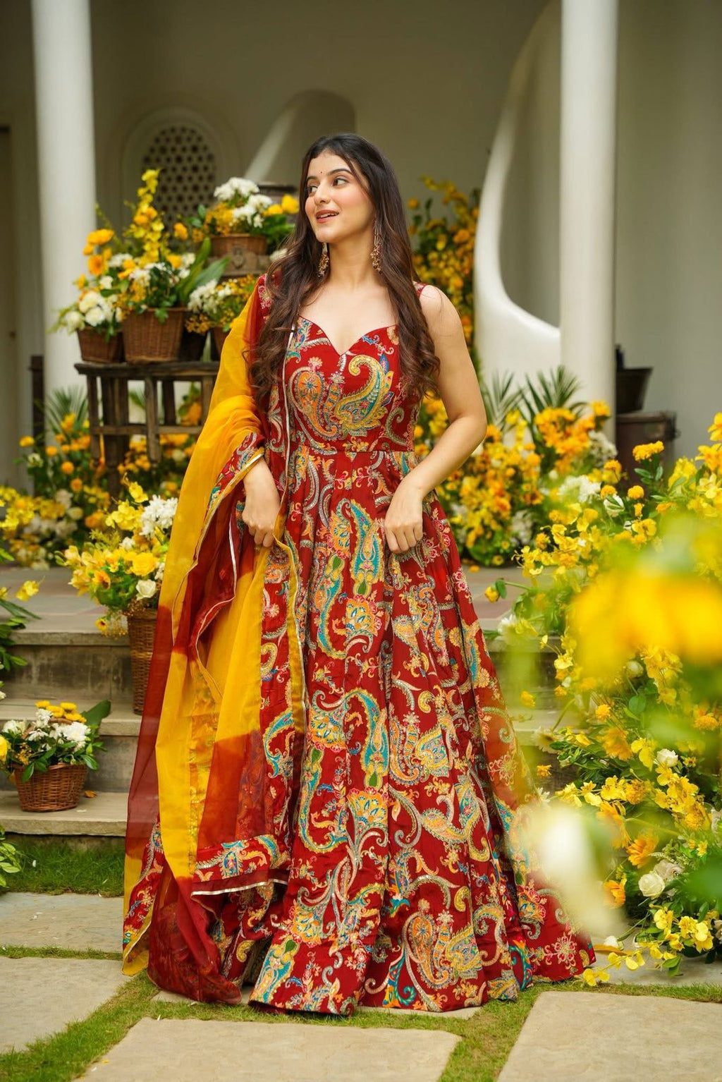Woman in a red and yellow traditional Anarkali Gown outfit standing in a garden with flowers and columns.