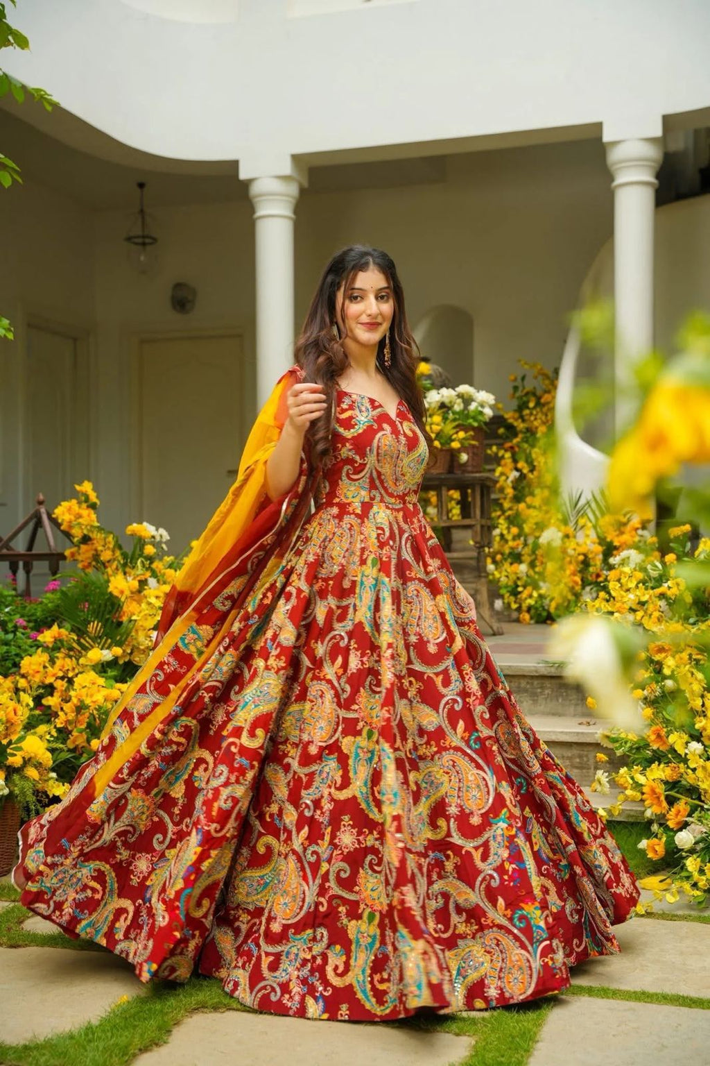 Woman in a red and multicolor patterned Anarkali Gown standing outdoors with flowers and columns in the background
