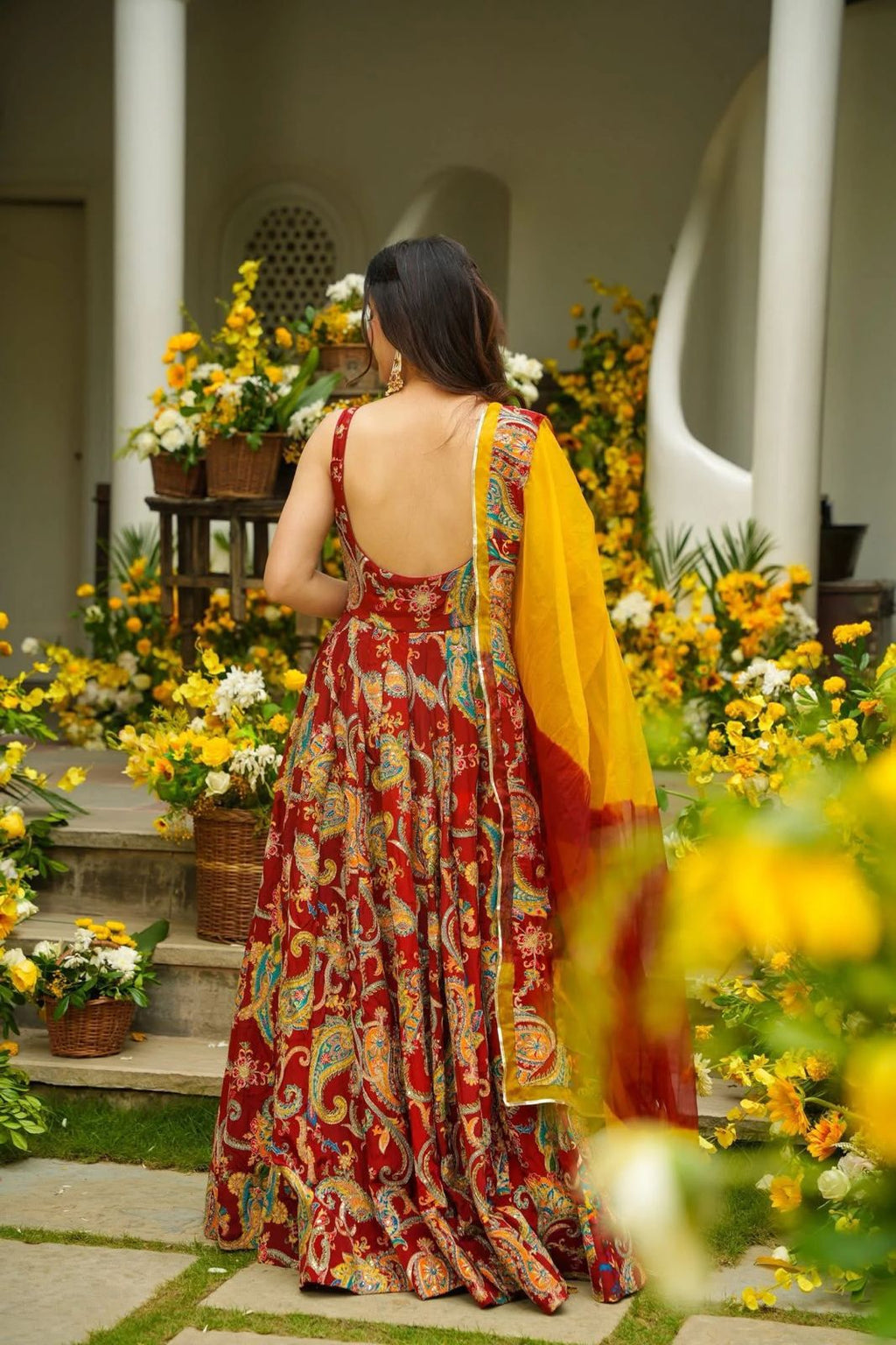 Woman in a colorful Anarkali Gown standing in a garden with flowers and architectural elements.