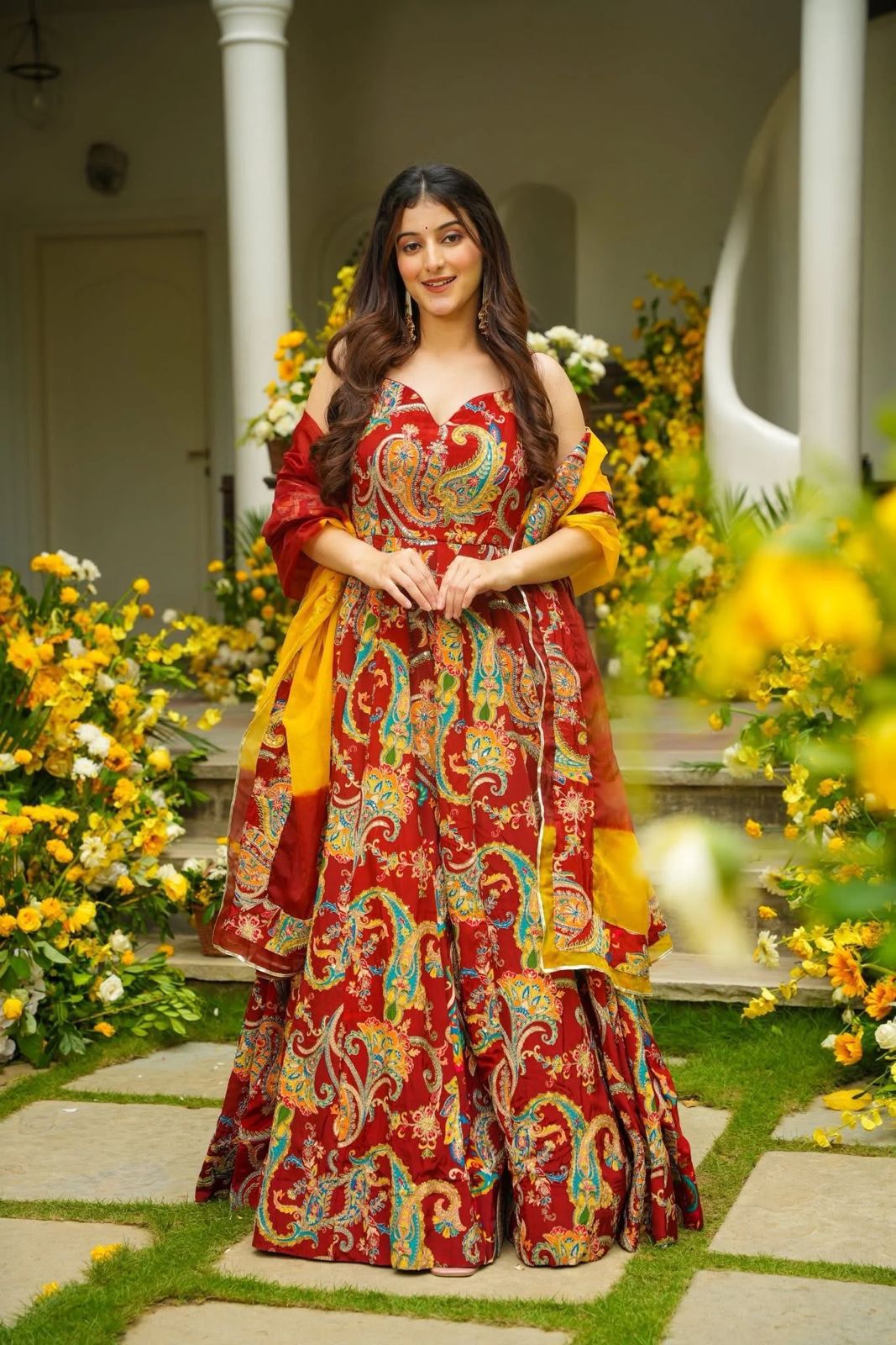 Woman in a colorful Anarkali Gown outfit standing in a garden with flowers and a building in the background