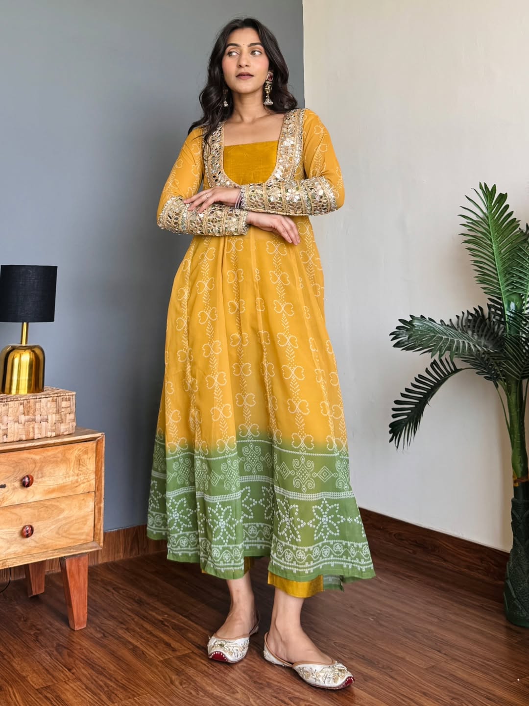 Woman wearing a yellow and green traditional Anarkali Kurta Set outfit in a room with a lamp and plant.