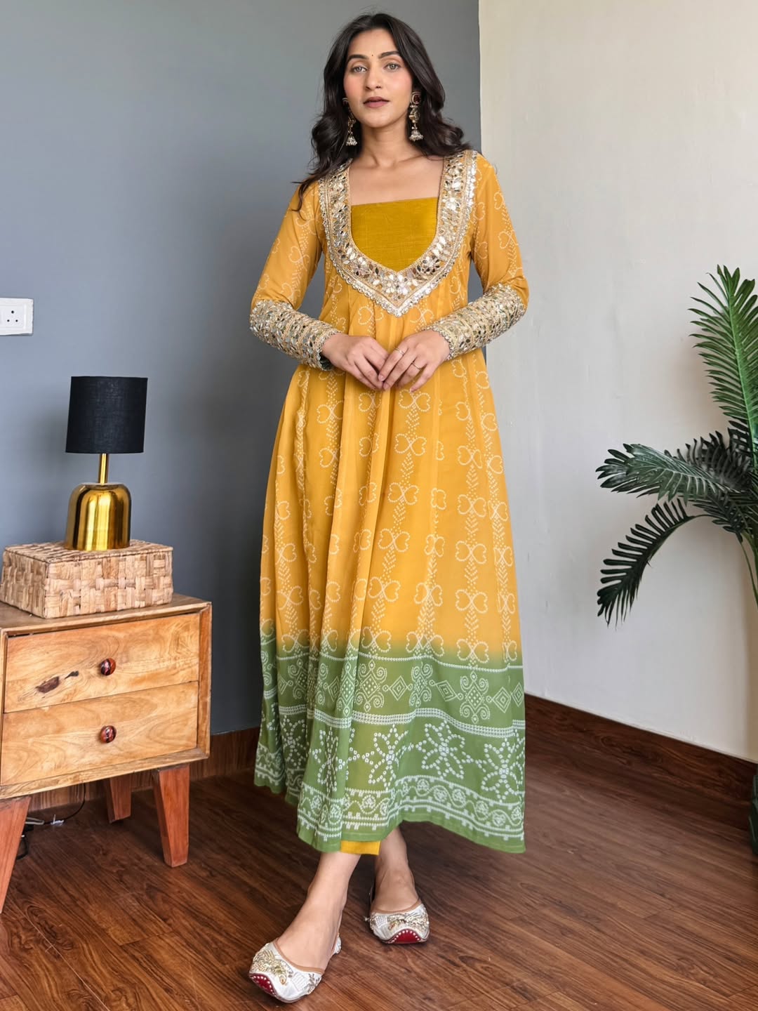 Woman wearing a mustard yellow and green traditional Anarkali Kurta Set outfit in a room with wooden furniture and a plant.