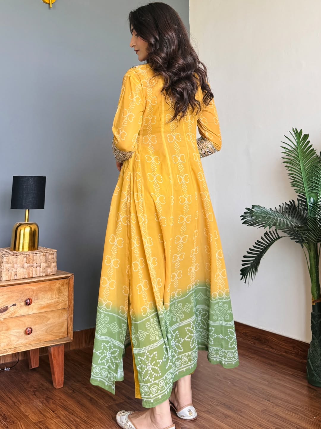 Woman wearing a yellow and green traditional Anarkali Kurta Set outfit in a room with a wooden dresser and plant.