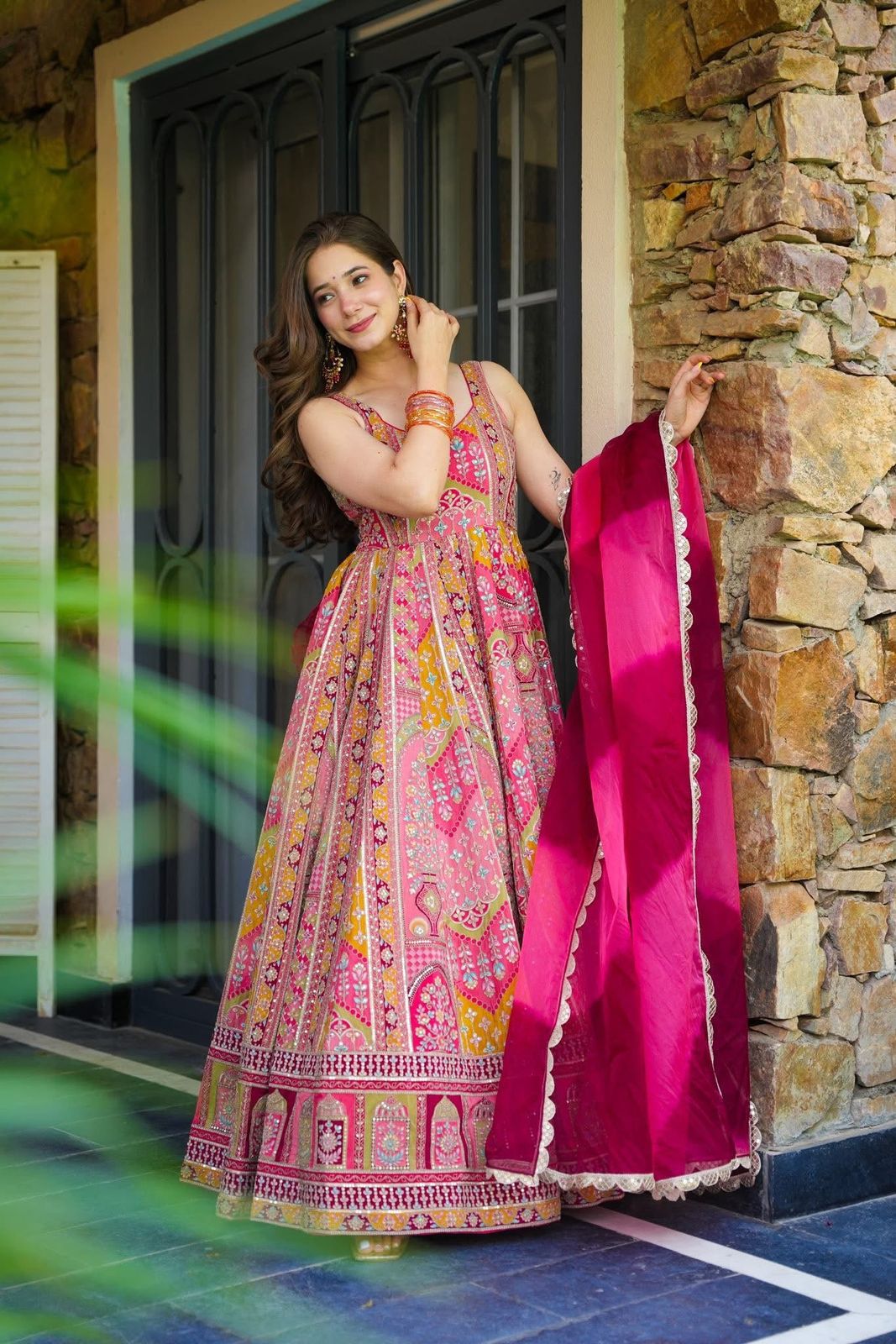 Woman in a colorful traditional Anarkali Gown outfit standing against a stone wall.