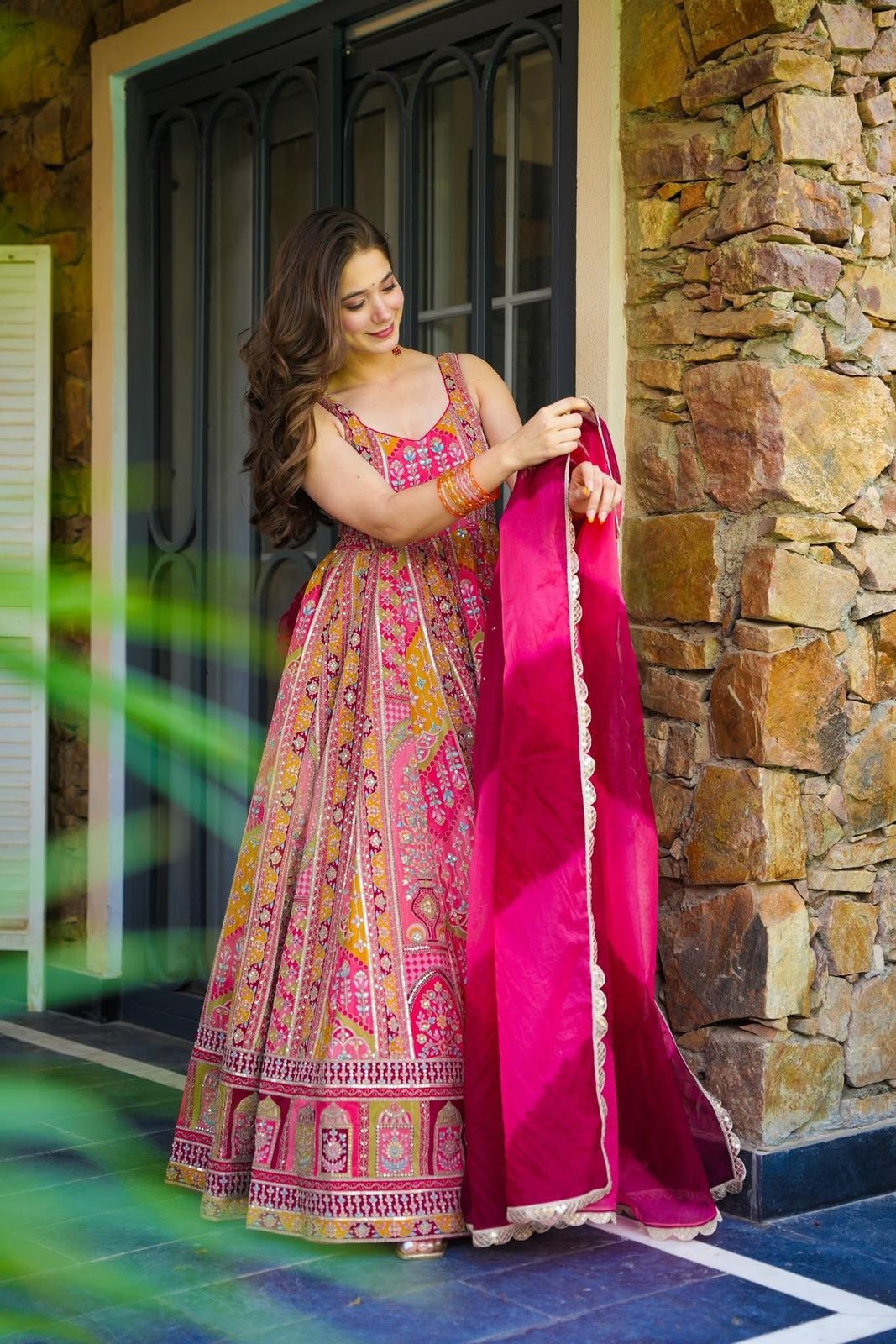 Woman in a colorful traditional Anarkali Gown outfit holding a pink dupatta against a stone wall.