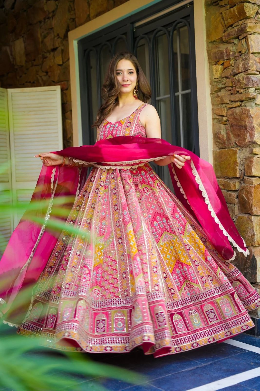 Woman in a colorful traditional Anarkali Gown outfit holding a pink dupatta, standing in front of a stone wall.