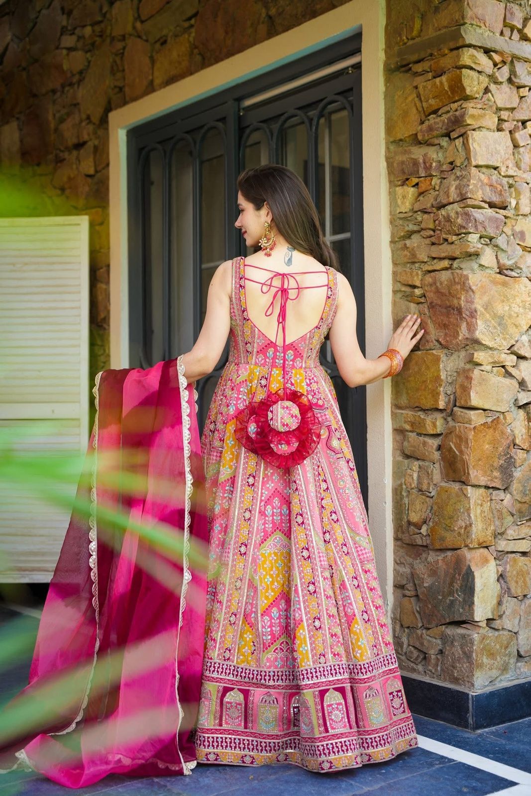 Woman in a pink and yellow patterned Anarkali Gown outfit with a floral accessory, standing in front of a stone wall.