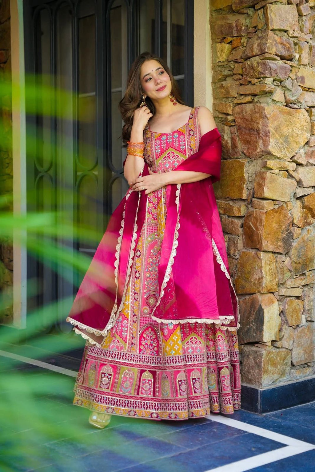 Woman in a colorful traditional Anarkali Gown outfit standing against a stone wall.