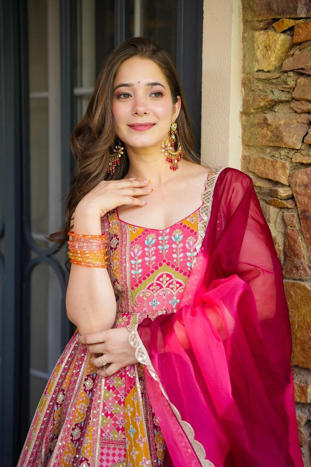 Woman in a colorful traditional Anarkali Gown outfit standing against a stone wall.
