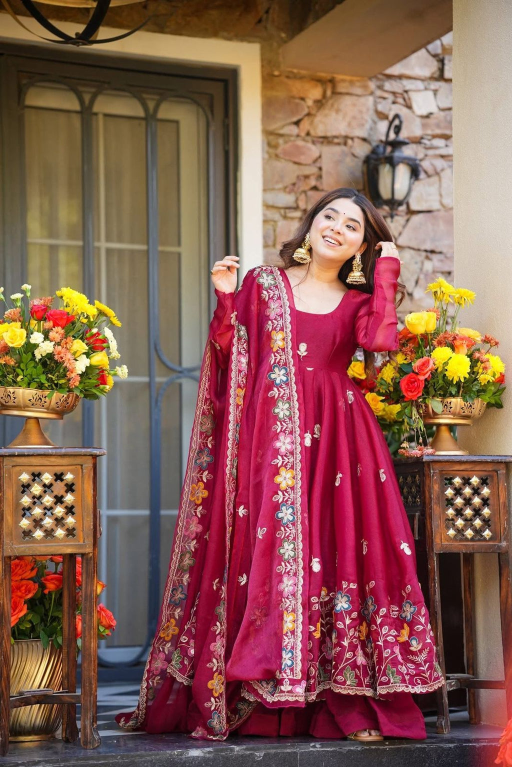 Woman in a red embroidered traditional Anarkali Palazzo Set outfit standing in front of a stone wall with floral decorations.