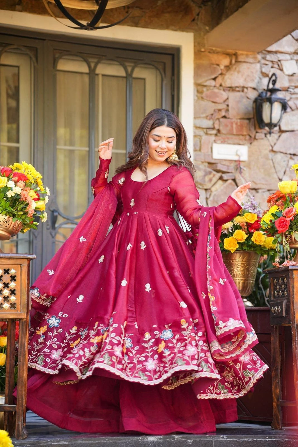 Woman in a red embroidered Anarkali Palazzo Set standing in front of a stone wall with floral decorations.