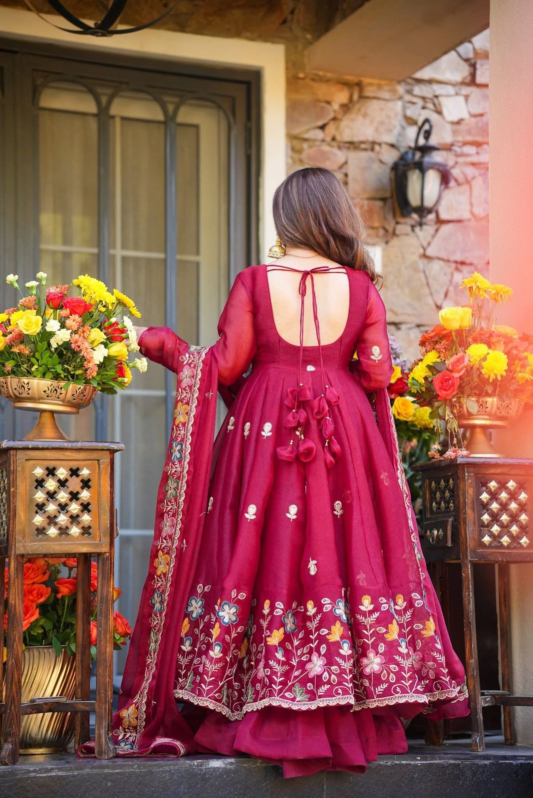 Woman in a red embroidered traditional Anarkali Palazzo Set outfit standing in front of a stone wall with floral decorations.