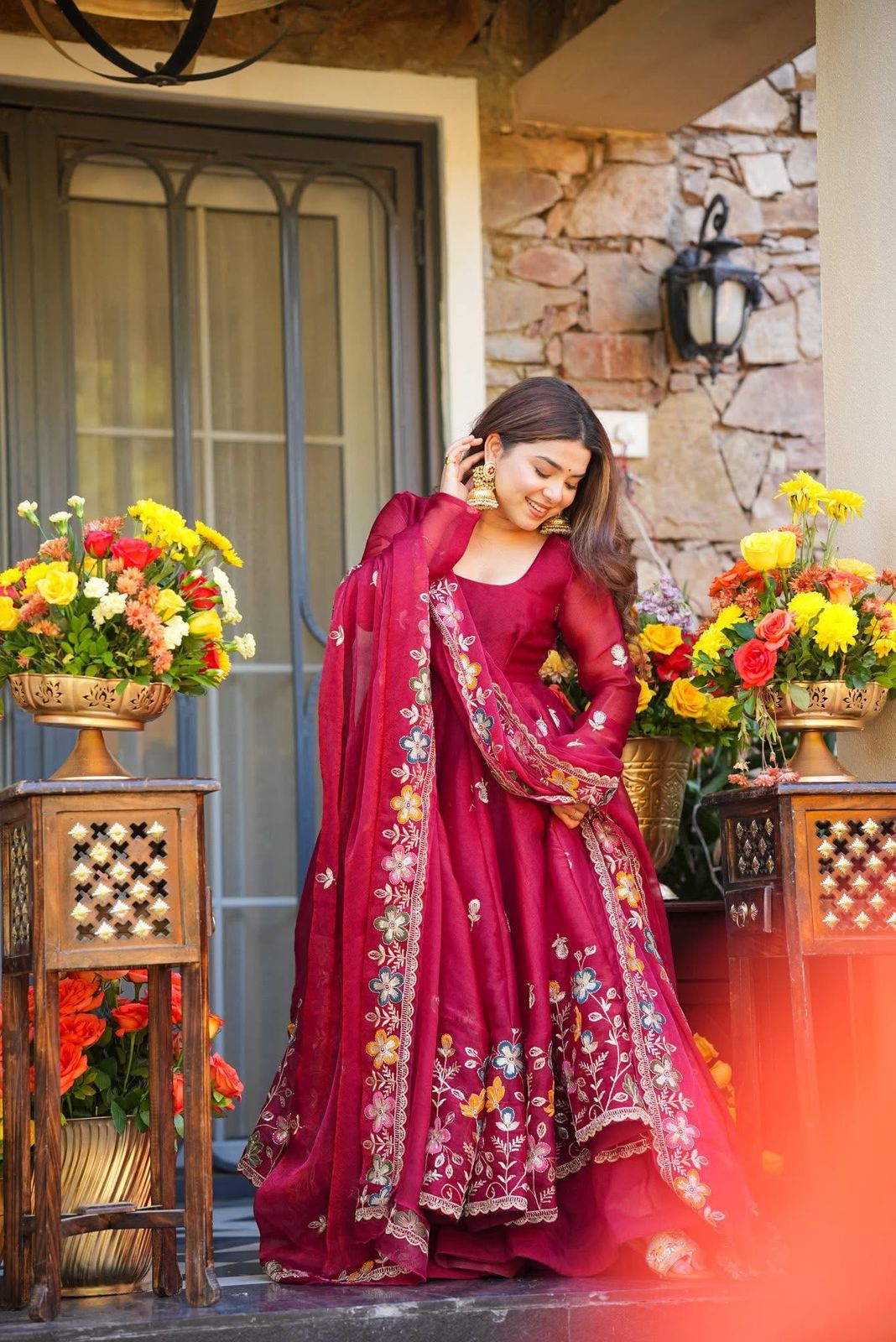 Woman in a red traditional Anarkali Palazzo Set outfit standing outdoors with floral arrangements.