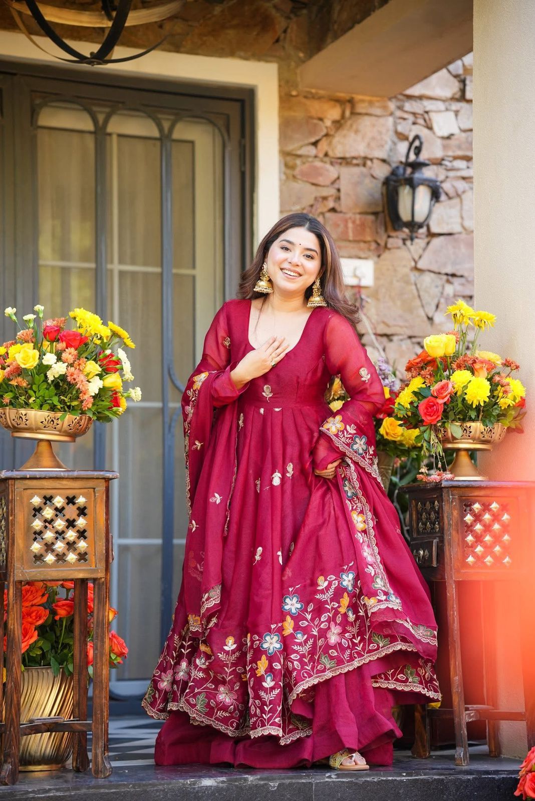 Woman in a red embroidered traditional Anarkali Palazzo Set outfit standing in front of a stone wall with floral arrangements.
