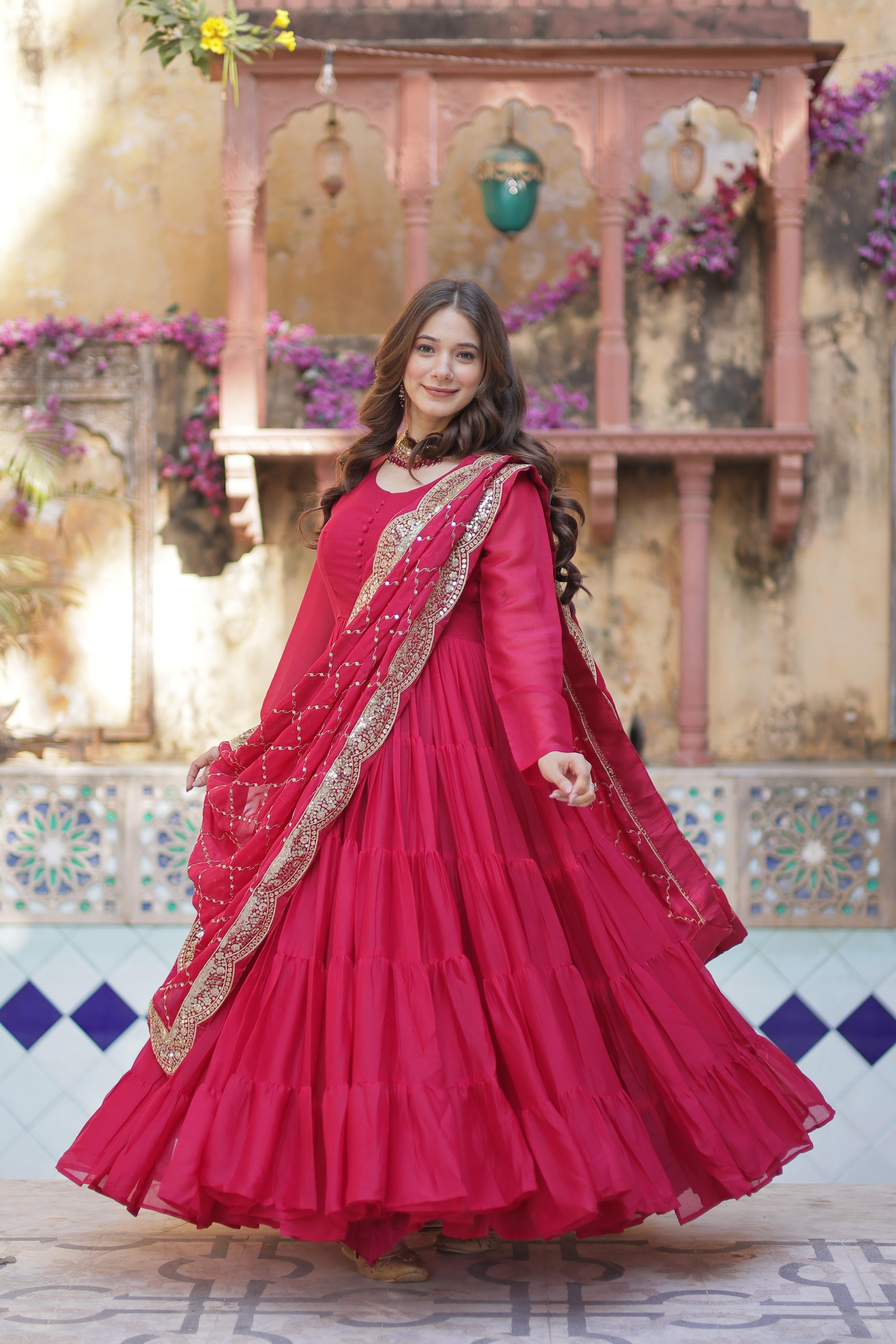 A woman in a red anarkali gown or dress with gold details standing in front of a decorative wall.
