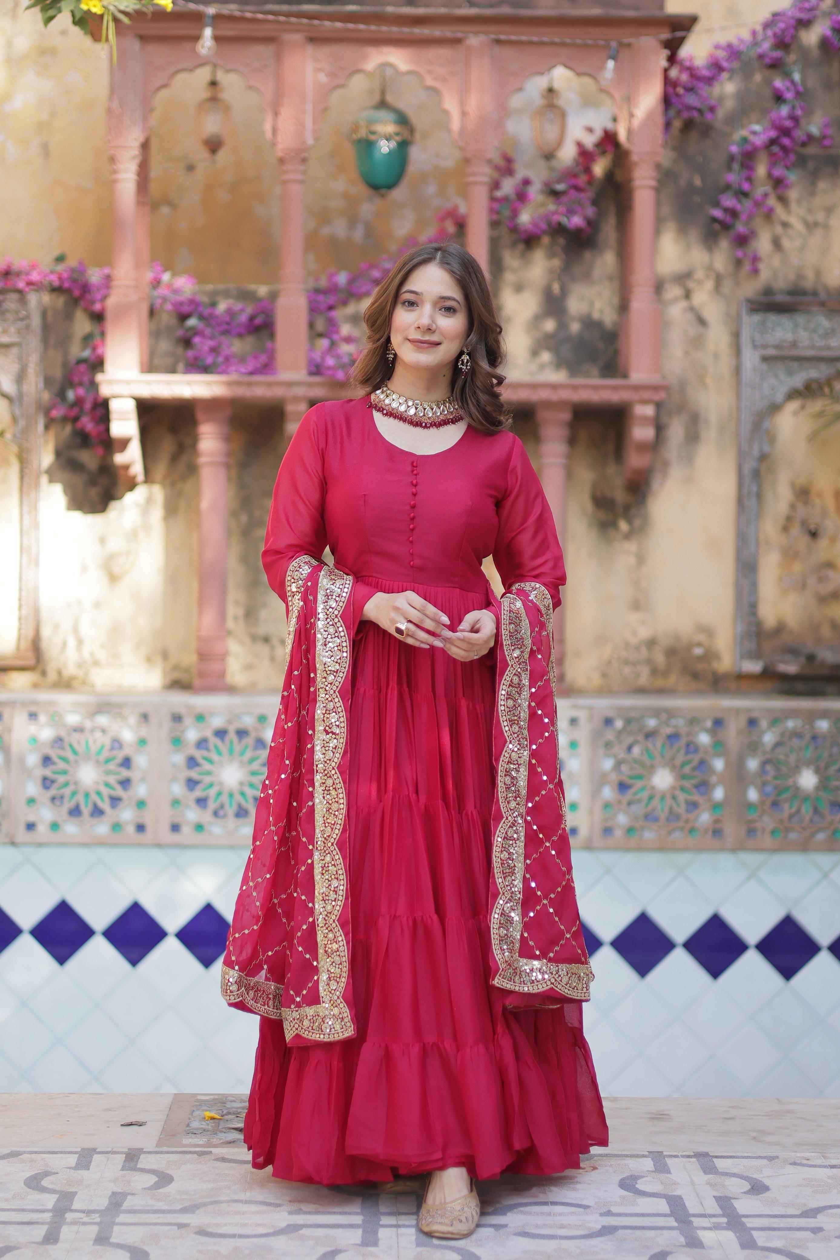A woman in a red anarkali gown or dress standing in front of decorative wall with flowers.