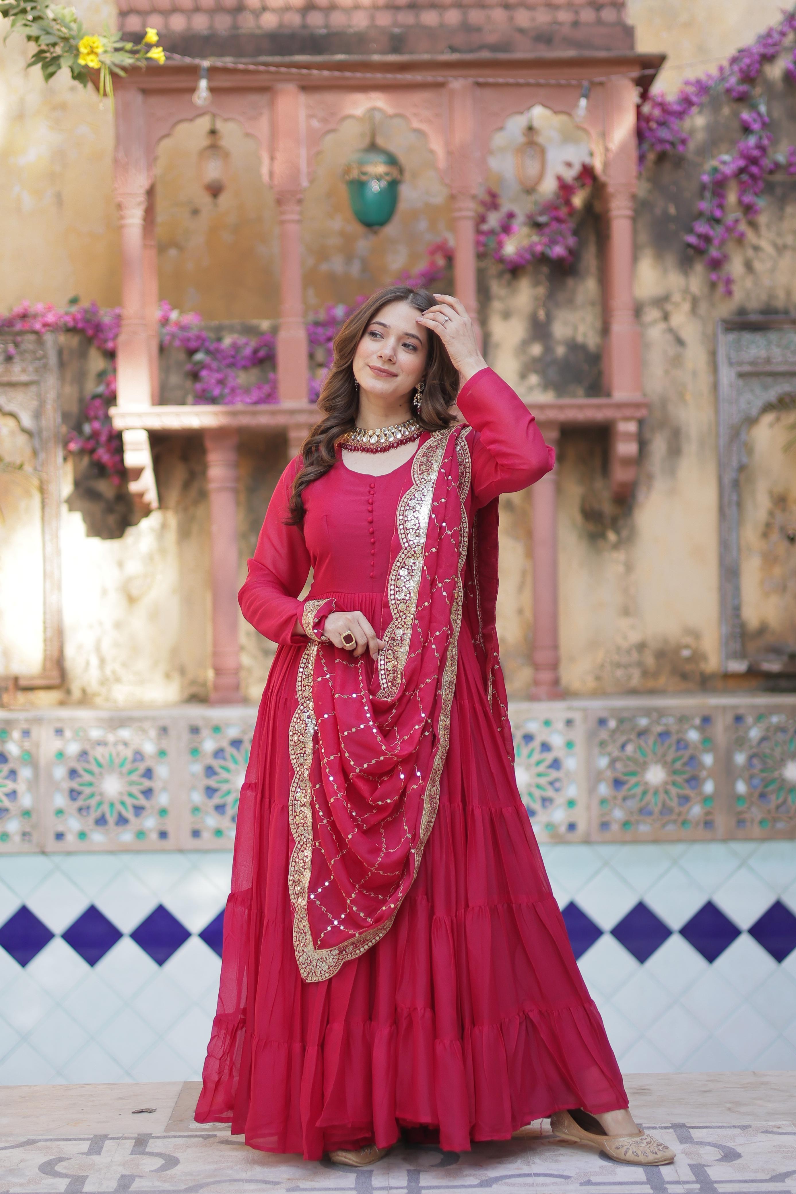 A woman in a red anarkali gown or dress standing in front of a decorative wall with flowers.