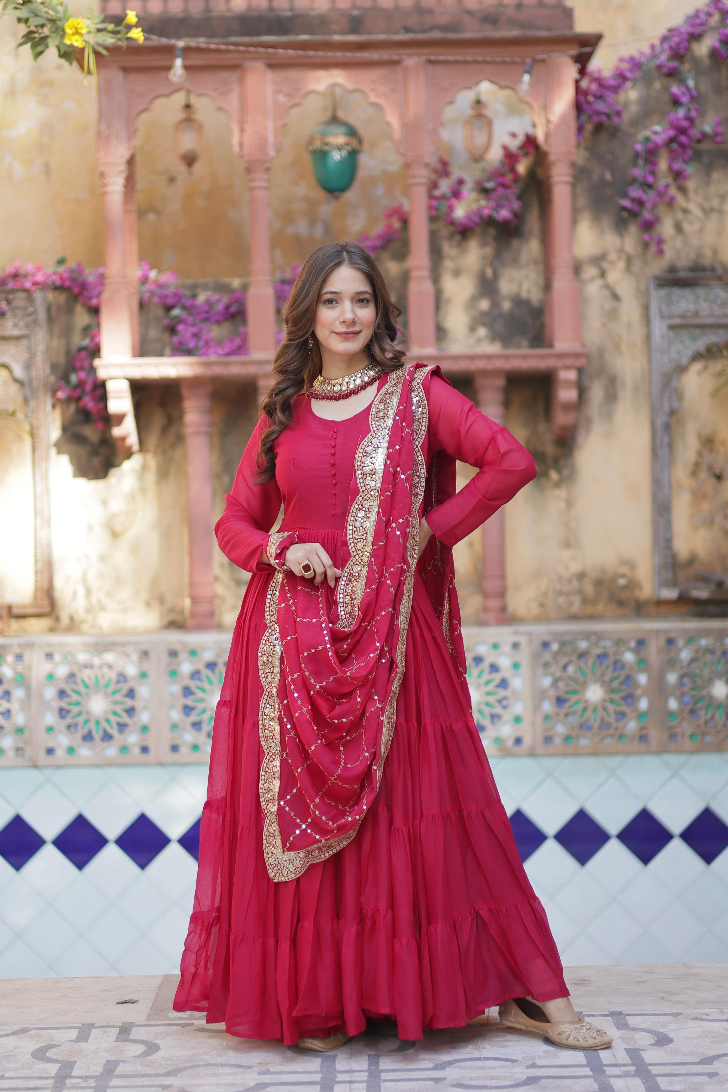A woman in a red anarkali gown or dress outfit with gold details standing in front of a decorative building.