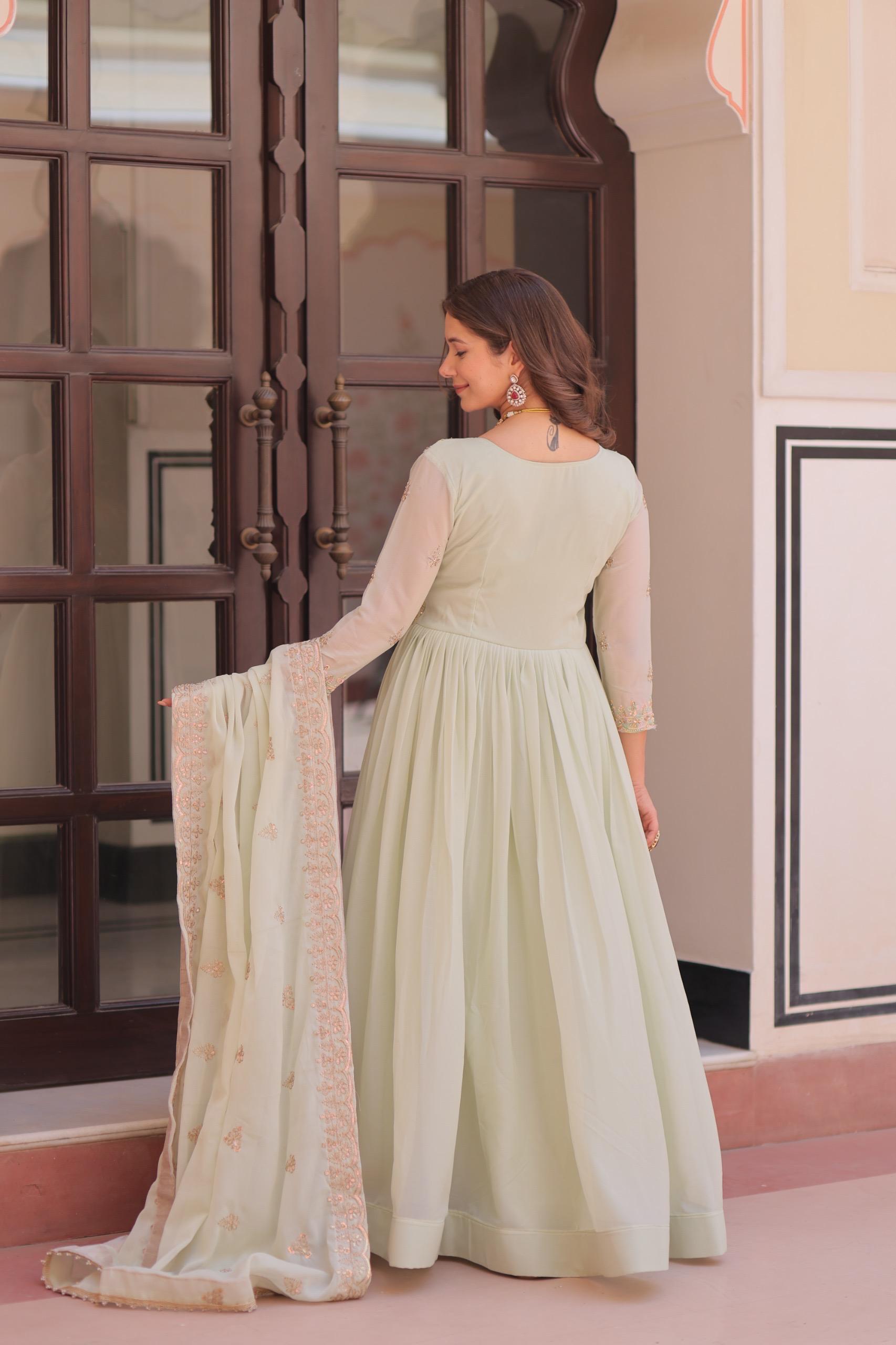 A woman in a light green Anarkali gown or dress holding a dupatta in an indoor setting with decorative elements.