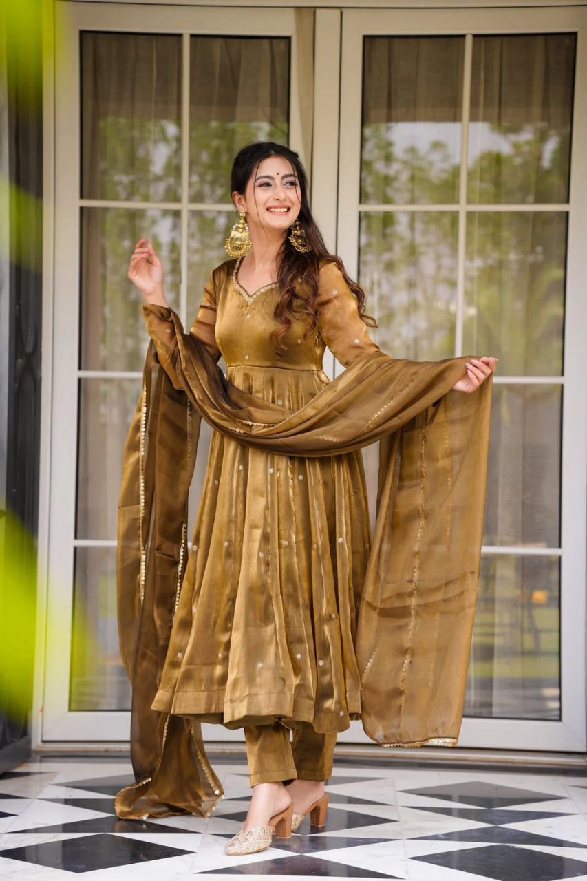 A woman in a gold Anarkali gown or dress standing in front of a glass door.