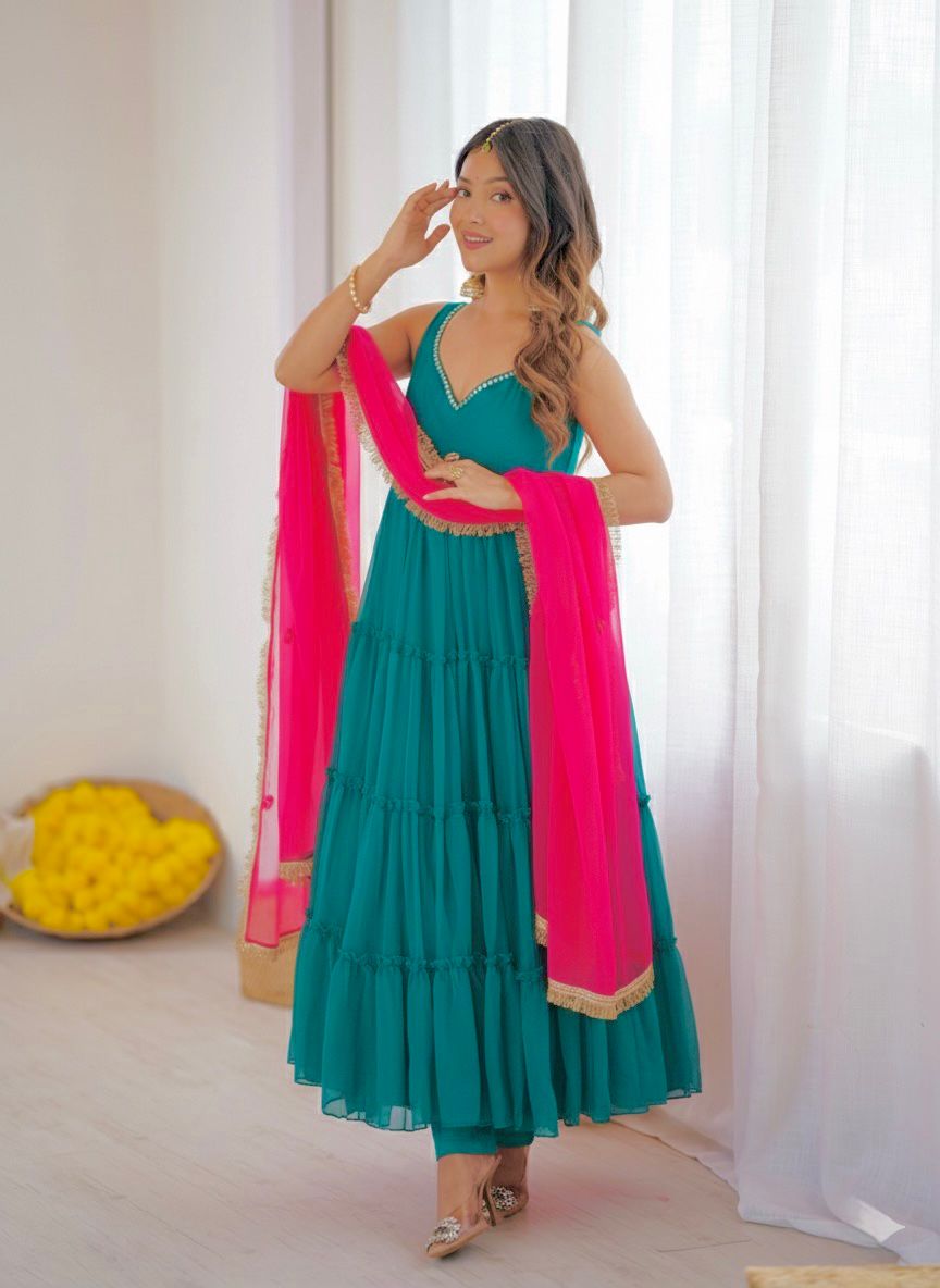 Woman in a blue anarkali dress with a pink dupatta standing against a white curtain.