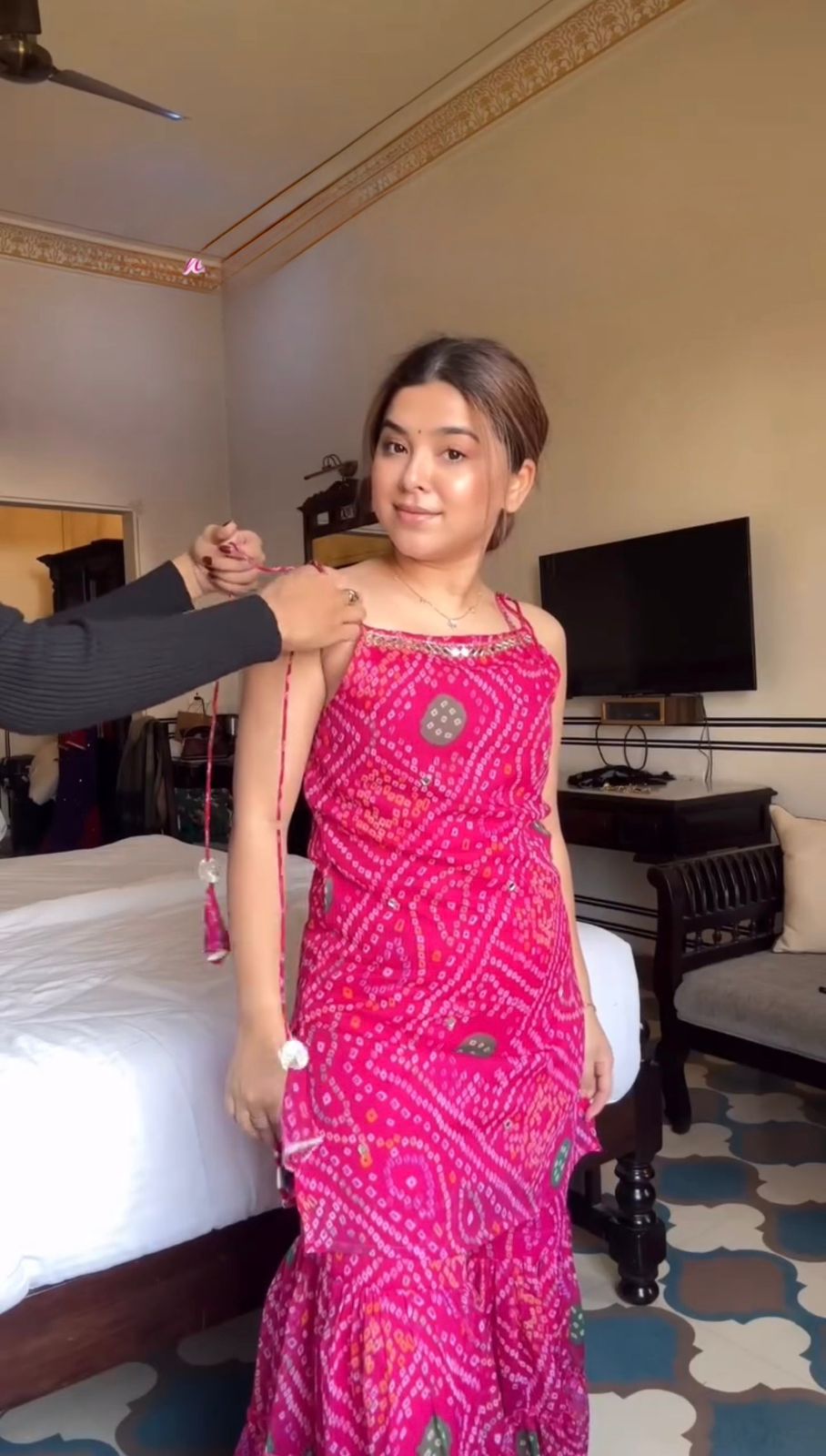 Woman in a pink sharara set being measured by an armorer in a room with furniture and decor.