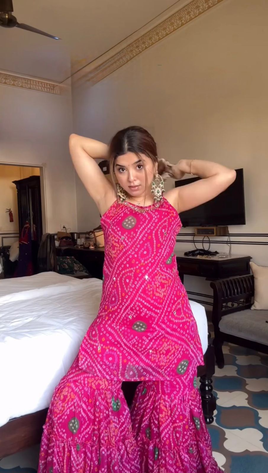 Woman in a pink sharara set traditional outfit posing in a room with a bed and television.