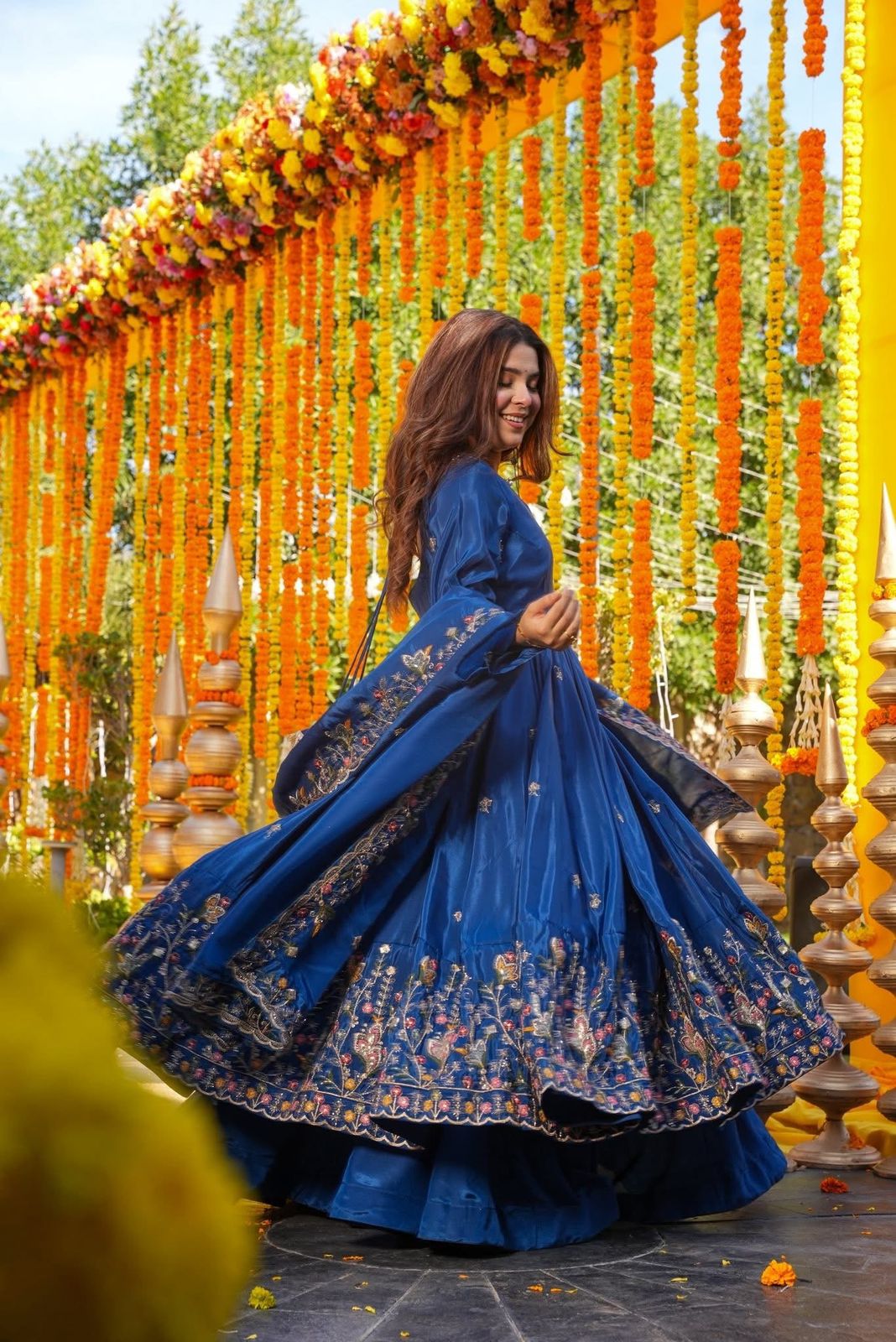 Woman in a blue embroidered traditional outfit standing in front of a decorative arch with flowers and lights.