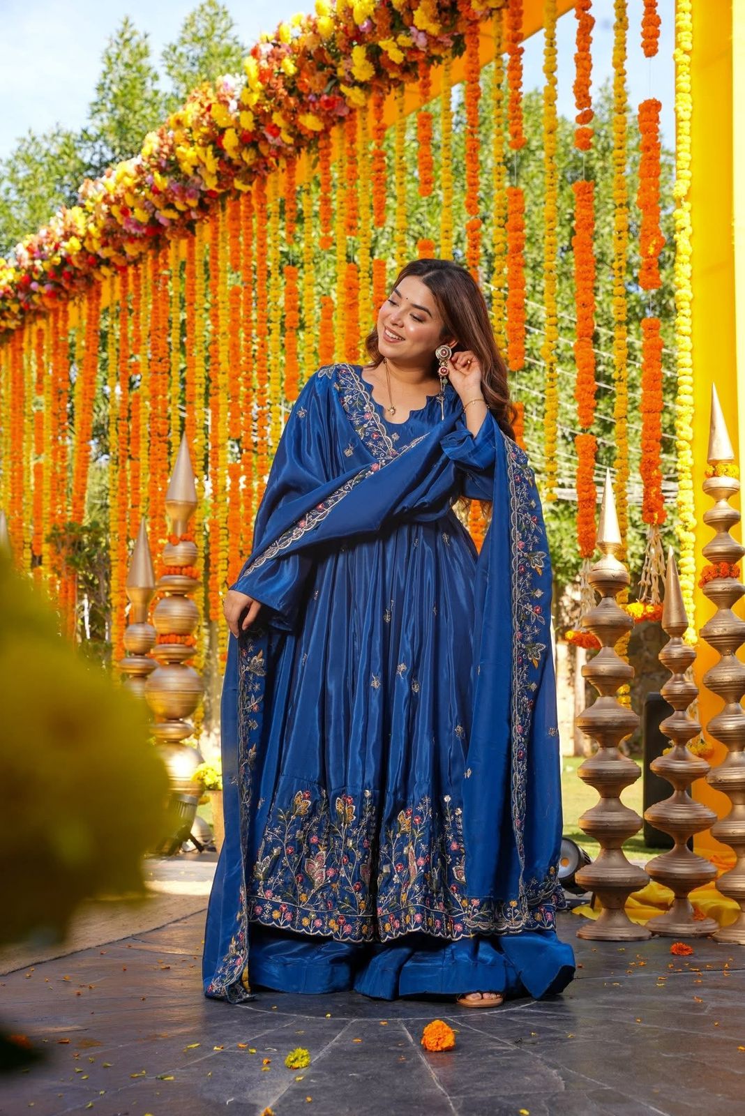 Woman in a blue traditional outfit standing in front of a decorated archway with flowers and lights.