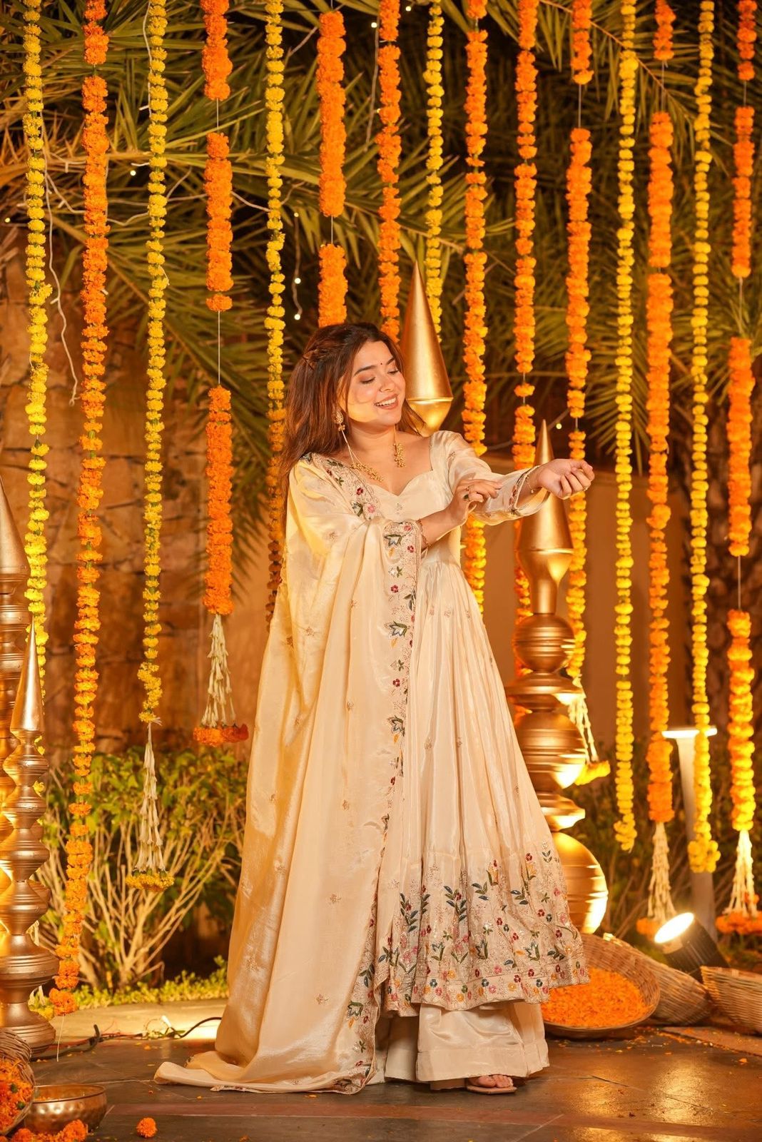 Woman in a traditional outfit standing in front of decorative floral and string lights.