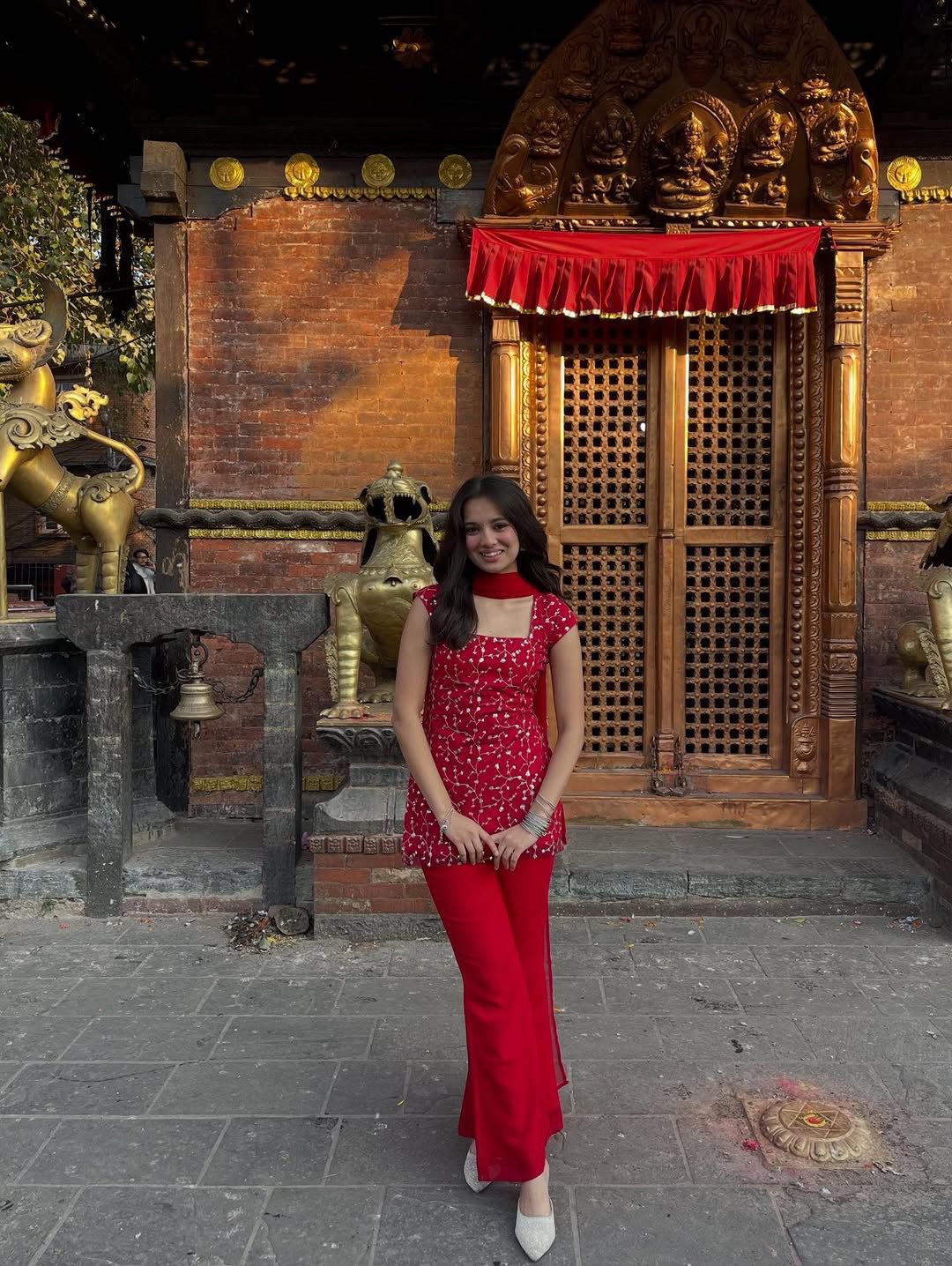 Woman in a red outfit standing in front of a wooden temple structure with intricate carvings.