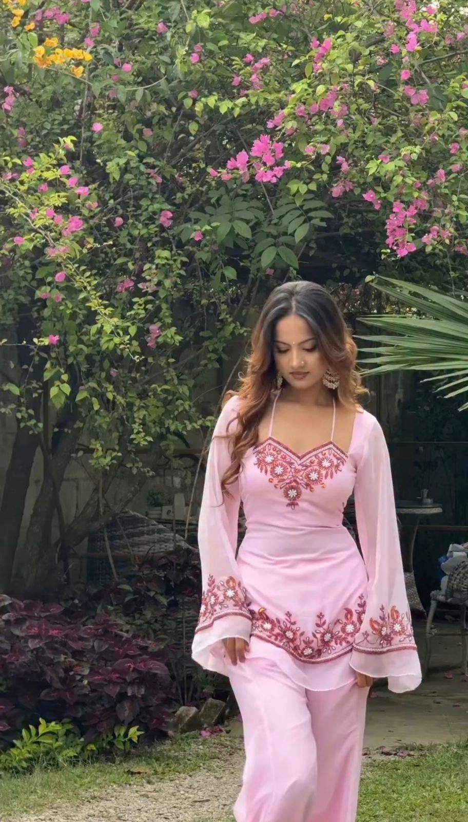 Woman in a pink traditional outfit standing in a garden with greenery and flowers.