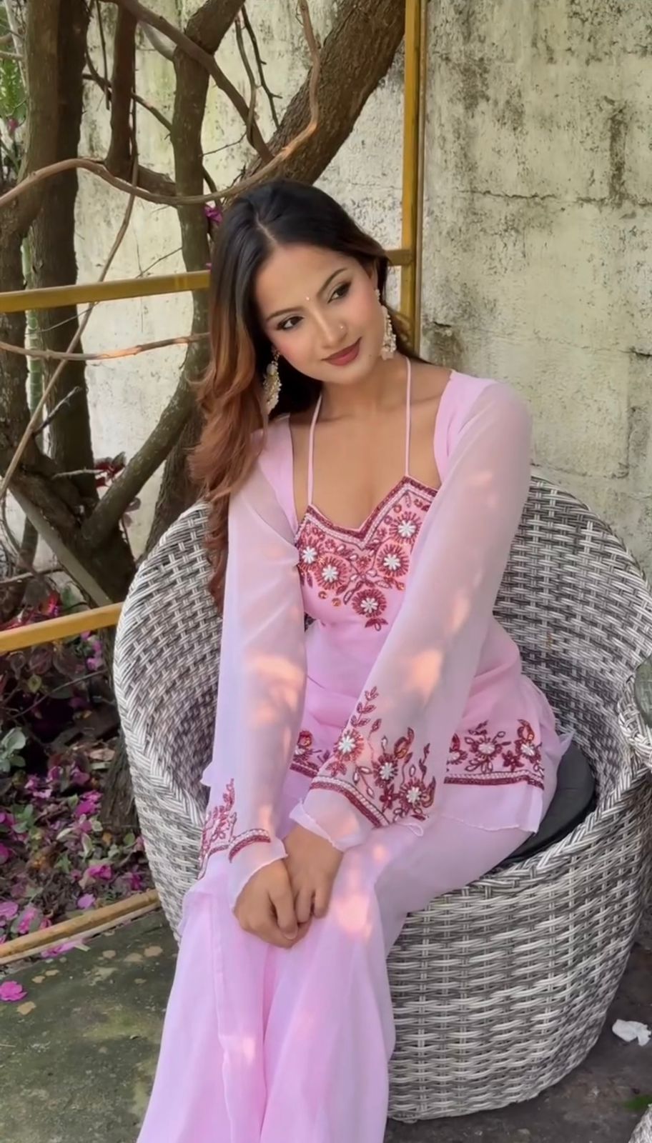 Woman in a pink traditional outfit sitting on a wicker chair outdoors.