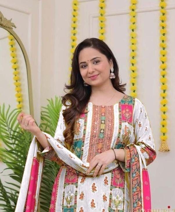 Woman in a traditional outfit with colorful patterns standing in front of decorative strings and a mirror.