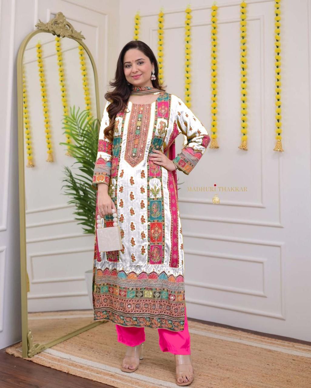 Woman in a colorful traditional outfit standing in a decorated room with a mirror and decorative elements.